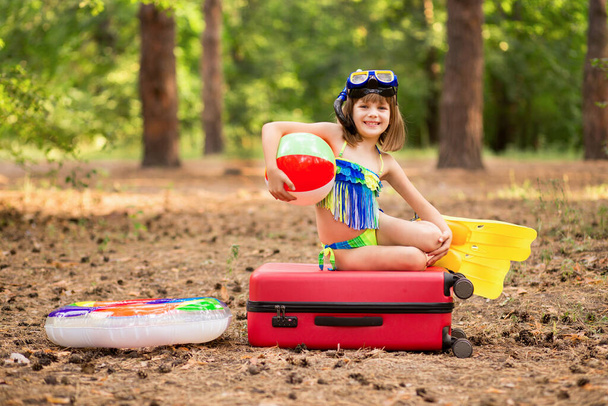 Энтузиастичная маленькая девочка в купальнике, плавниках и маске с круговым и плавательным мячом, упакованная в чемодан для летних каникул - Фото, изображение