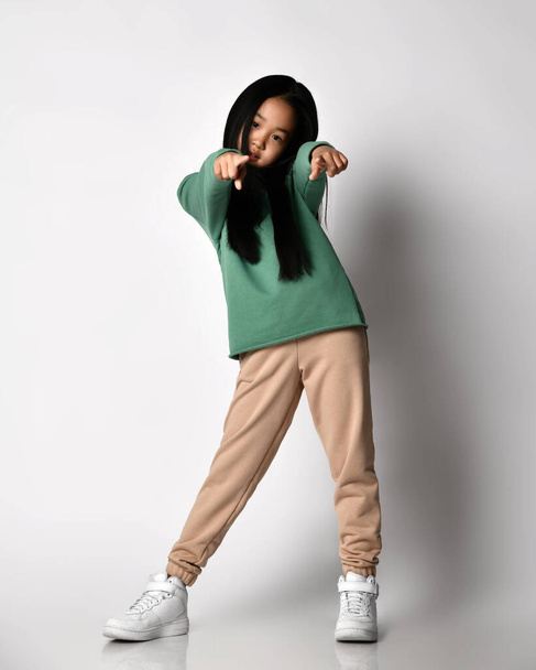 Asian kid girl in sneakers, green pants and hoodie, sweater stands pointing with fingers of both hands at camera - Фото, зображення