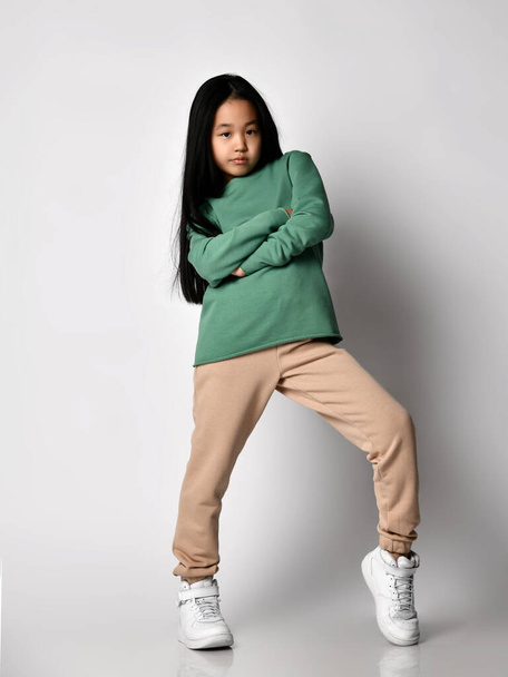 Cool Asian kid girl green sportswear pants, hoodie, sweater and sneakers stands holding arms crossed at chest - Foto, Bild