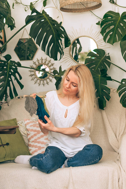 A young blonde woman weaving macrame in a home workshop.There are many indoor plants in the room.Handmade concept. - Fotoğraf, Görsel