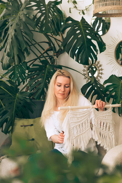 A young blonde woman weaving macrame in a home workshop.There are many indoor plants in the room.Handmade concept. - Foto, imagen