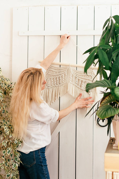 Young attractive blonde woman hanging a white macrame decor on the wall.Indoor plants in the background.Handmade concept.Selective focus,rear view. - Foto, imagen