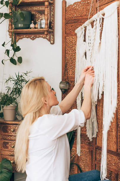 A young attractive blonde woman in a white blouse and jeans is weaving macrame.The home decor hangs on a patterned wooden screen.The interior of the workshop is decorated with indoor plants,side view. - Photo, image