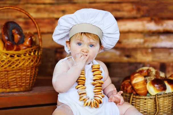 baby boy in a chefs hat and apron with a bunch of bagels around his neck sits against the background of bakery products in a bread shop. - Foto, afbeelding