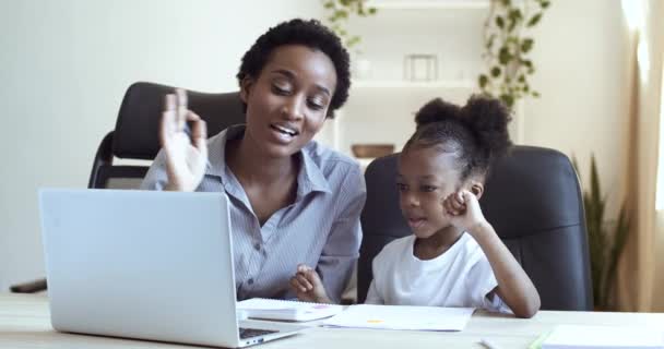 アフリカ系アメリカ人の母親と娘の女子高生の黒人の子供がノートパソコンのテーブルに座っていると、手を振って笑う家族とインターネットでリモート通信します。子供はオンラインで宿題を笑っている - 映像、動画