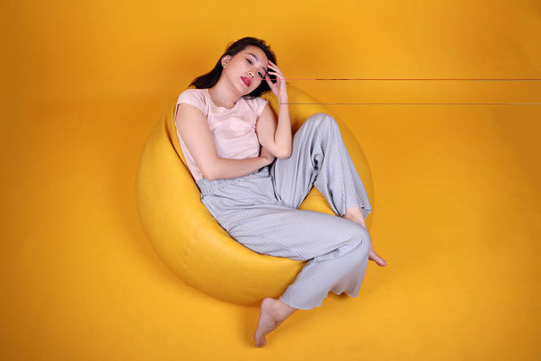 Beautiful young south east Asian woman sits on a yellow beanbag seat orange yellow color background pose fashion style elegant beauty mood expression rest relax think emotion sleep top view - Fotó, kép