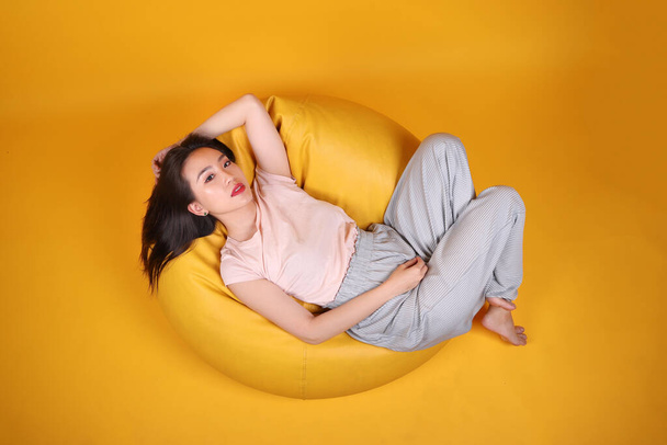Schöne junge südostasiatische Frau sitzt auf einem gelben Sitzsack Sitz orange gelb Farbe Hintergrund Pose Mode Stil elegante Schönheit Stimmung Ausdruck Ruhe denken Emotionen Schlaf Draufsicht - Foto, Bild