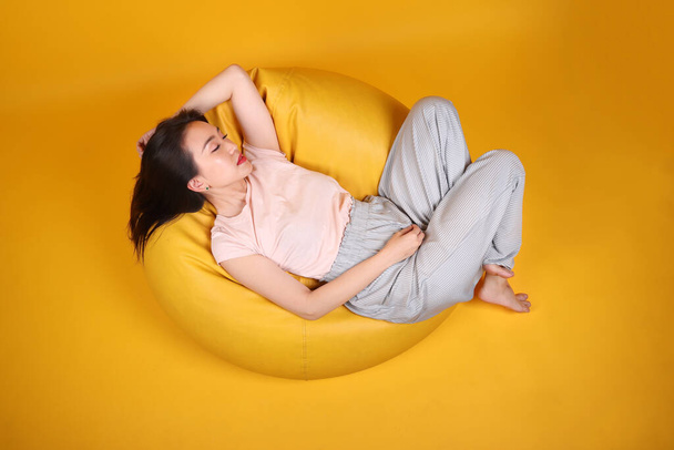 Όμορφη νεαρή νοτιοανατολική ασιατική γυναίκα κάθεται σε ένα κίτρινο κάθισμα beanbag πορτοκαλί χρώμα φόντο θέτουν στυλ μόδας κομψή ομορφιά διάθεση έκφραση ξεκουραστείτε σκεφτείτε συναίσθημα ύπνο κορυφή άποψη - Φωτογραφία, εικόνα