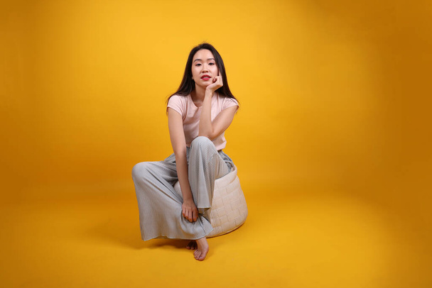 Schöne junge südostasiatische Frau sitzt auf einem weißen Sitzsack Sitz orange gelb Farbe Hintergrund Pose Mode Stil elegante Schönheit Stimmungsausdruck entspannen Lächeln aussehen - Foto, Bild