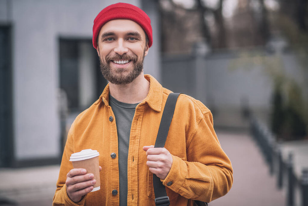 Man in a red hat and orange jacket with a coffee cup in hands - Fotografie, Obrázek