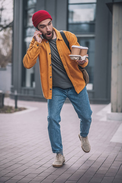 Young bearded man in a red hat talking on the phone and dropping the coffee cups - Foto, Bild