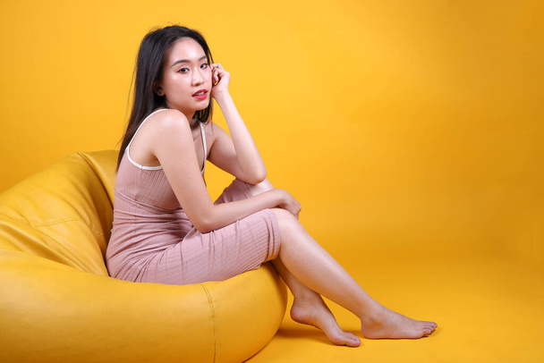 Beautiful young south east Asian woman sit on a yellow orange beanbag seat color background pose fashion style elegant beauty mood - Photo, Image