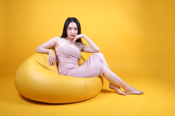 Beautiful young south east Asian woman sit on a yellow orange beanbag seat color background pose fashion style elegant beauty mood - Foto, Imagem