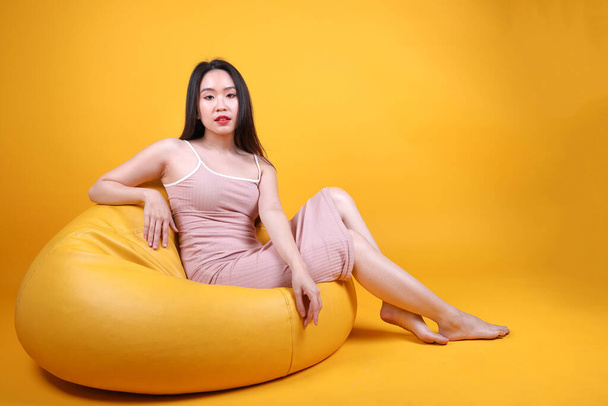 Beautiful young south east Asian woman sit on a yellow orange beanbag seat color background pose fashion style elegant beauty mood - Photo, Image