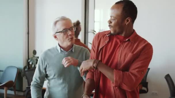Veselý starší běloch se usmívá, zdraví mladého afroamerického spolupracovníka s hrboly pěstí a klábosí s ním, zatímco se setkávají v úřadu na začátku pracovního dne - Záběry, video