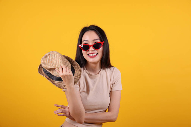 Güzel, genç, Güneydoğu Asyalı kadın kırmızı çerçeveli güneş gözlüklü şapka moda zirvesi sarı turuncu arka planda elinde şapkayla mutlu bir gülümseme. - Fotoğraf, Görsel