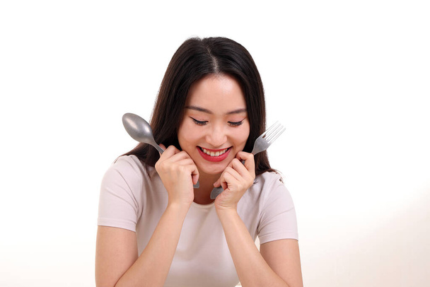 Όμορφη νεαρή νοτιοανατολική ασιατική γυναίκα προσποιούνται ενεργεί κρατώντας άδειο κουτάλι πιρούνι στο χέρι τρώνε γεύση δείτε λευκό πιάτο στο τραπέζι λευκό φόντο χαμόγελο ευτυχισμένη έξοδο - Φωτογραφία, εικόνα