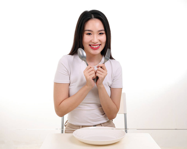 Mooie jonge Zuidoost-Aziatische vrouw doen alsof poseren met lege vork lepel in de hand eten smaak look zie witte plaat op tafel witte achtergrond glimlach gelukkig - Foto, afbeelding