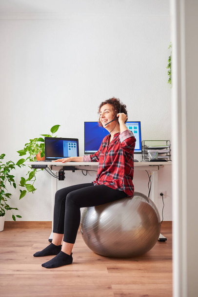 Woman teleworking sitting on a fitball in front of her desk talking on a headset - Foto, imagen