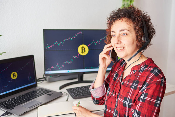 Trader femminile opera con bitcoin dalla sua scrivania in piedi mentre parla sul telefono cellulare e indossa un auricolare - Foto, immagini