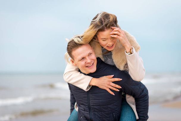 Szczęśliwy mężczyzna podwozi swoją kobietę na barana i śmieje się na plaży. Uśmiechnięty facet w miłości prowadzenia z powrotem jej dziewczyna i dobrze się bawić.Szczęśliwa zdrowa para na świeżym powietrzu korzystających z życia. - Zdjęcie, obraz