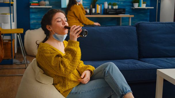 Relaxed woman taking off face mask looking at camera drinking beer - Photo, Image
