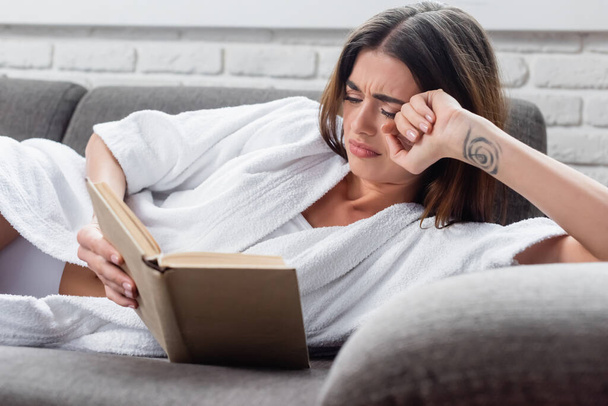 upset young adult woman reading book on grey couch at home - Foto, afbeelding