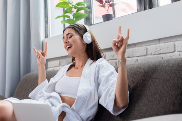 young adult woman with rock gesture sitting with crossed legs and listening music in headphones in living room - Фото, изображение