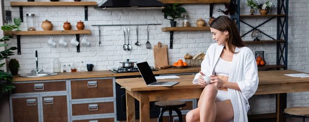 smiling young adult woman in white bathrobe sitting near laptop in loft, banner - Foto, imagen