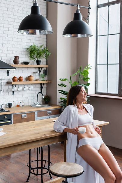 positive young adult woman in white bathrobe holding cup of coffee in modern loft - Φωτογραφία, εικόνα