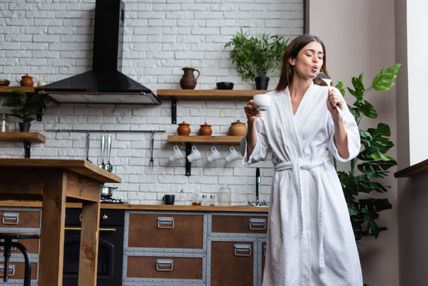 playful young adult woman in white bathrobe holding cup of coffee and spatula and singing in modern loft - Fotoğraf, Görsel