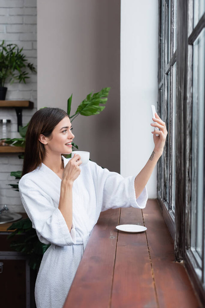 улыбающаяся молодая женщина в халате делает селфи на сотовом телефоне возле окна в современном лофте  - Фото, изображение