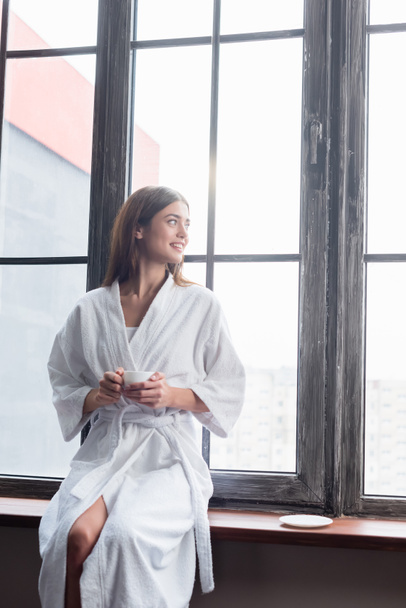 pretty young adult woman in bathrobe sitting with cup of coffee near window at home - Photo, Image