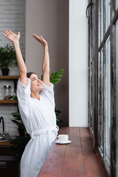 feliz joven mujer adulta en albornoz de pie cerca de la ventana con las manos en el aire en la sala de estar moderna - Foto, Imagen