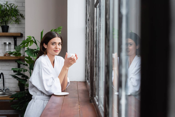 happy young adult woman holding cup of coffee near window with reflection in modern living room - Foto, Bild