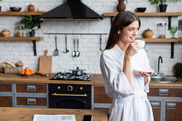 smiling young adult woman in bathrobe drinking coffee near table with newspaper and cellphone in modern loft - Foto, imagen