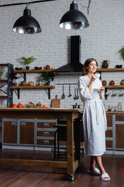 full length view of happy young adult woman in bathrobe drinking coffee in modern loft - Foto, afbeelding