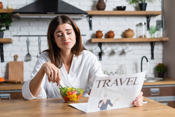entediado jovem mulher adulta em roupão de banho comer salada e ler jornal de viagem na cozinha moderna - Foto, Imagem