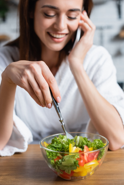 uśmiechnięta młoda kobieta w szlafroku je sałatkę i rozmawia przez telefon komórkowy w kuchni - Zdjęcie, obraz