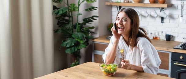 mladá dospělá žena v županu jíst salát a mluvit na mobilu v, banner - Fotografie, Obrázek