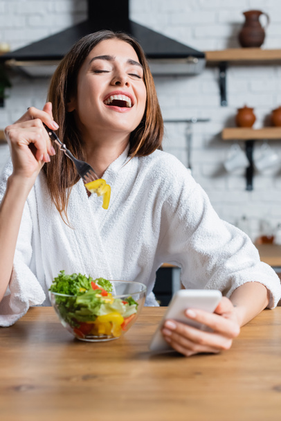 laughing young adult woman in bathrobe eating vegetables salad and using cellphone in modern kitchen - Fotografie, Obrázek