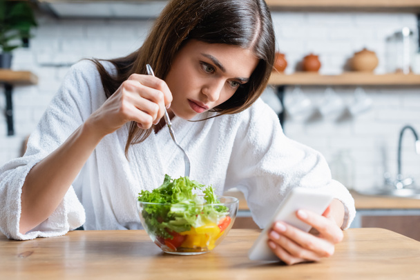 mujer adulta joven molesta en albornoz comer ensalada de verduras y el uso de teléfono celular en la cocina moderna - Foto, Imagen