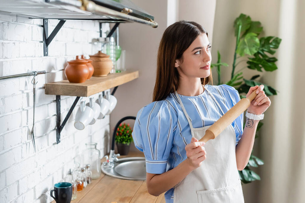 thoughtful young adult woman in apron standing with dough rolling pin in modern kitchen - Fotografie, Obrázek