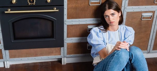upset young adult woman sitting on floor near oven with in modern kitchen, banner - Valokuva, kuva