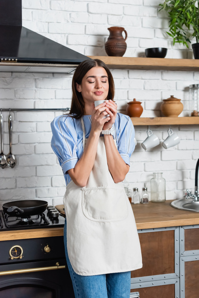 sonriente mujer joven adulta oliendo aroma de café en taza blanca en la cocina moderna - Foto, Imagen