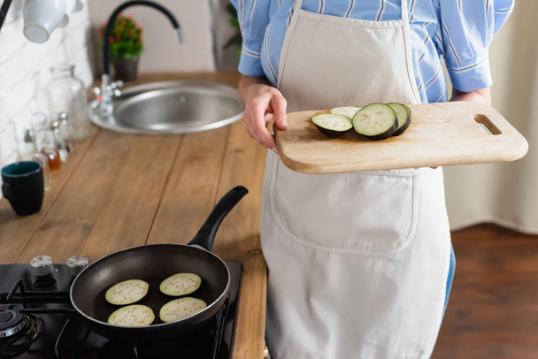 обрізаний вид молодої дорослої жінки, що тримає скибочки баклажанів на обробній дошці біля сковороди на кухні
 - Фото, зображення