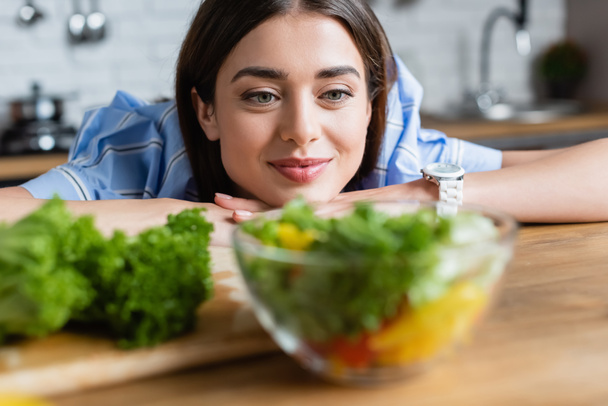 smiling young adult woman looking at prepared vegetables salad in kitchen - Fotó, kép
