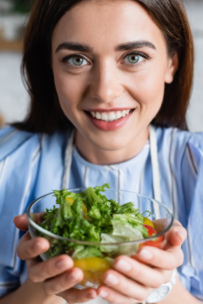 portrait of smiling young adult woman holding fresh vegetables salad in hands on blurred background - Photo, image