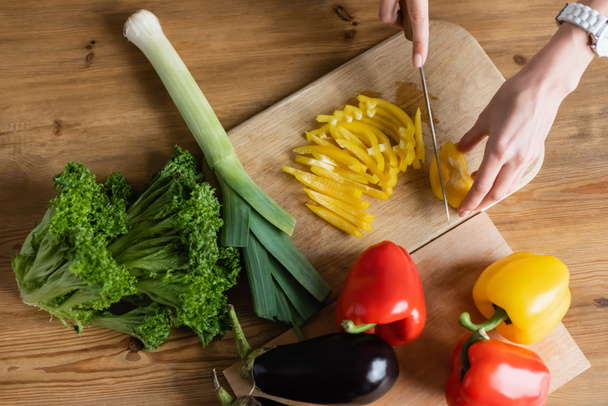 vista superior de las manos femeninas picando pimiento amarillo en la tabla de cortar con otras verduras en la mesa en la cocina - Foto, Imagen