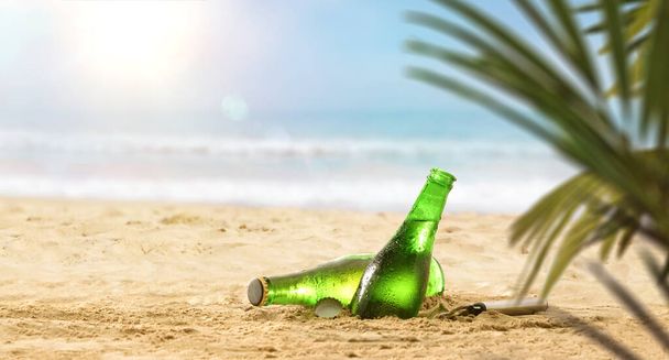 Detail of fresh beer bottles half buried in the sand on a beach with palm leaves on a hot day. Front view detail. - Fotografie, Obrázek